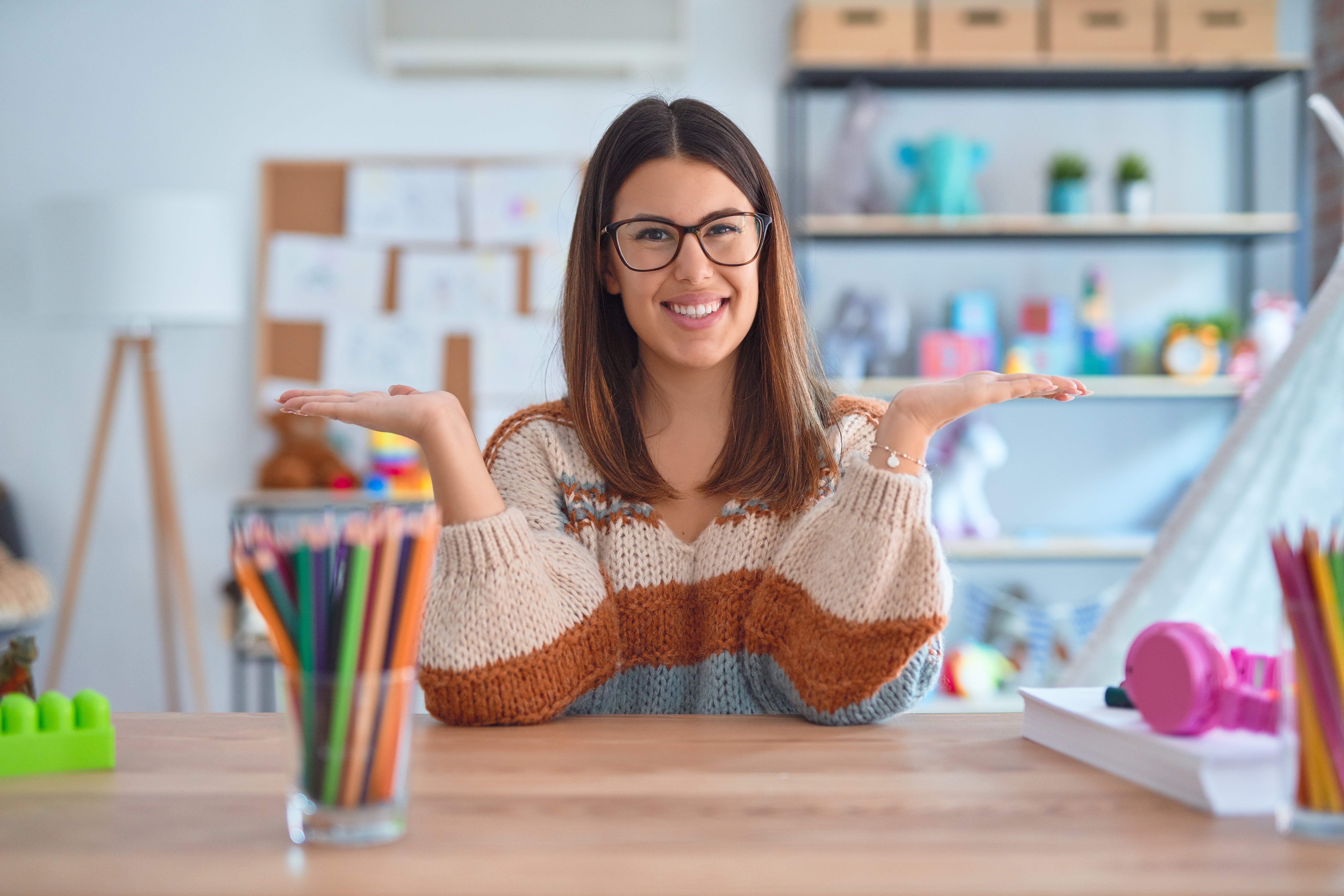 Young beautiful teacher woman wearing sweater and glasses sitting on desk at kindergarten smiling showing both hands open palms, presenting and advertising comparison and balance